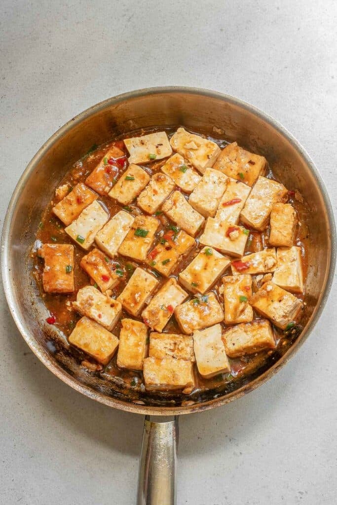 A pan filled with tofu cubes cooking in sauce with visible herbs and small pieces of vegetables.