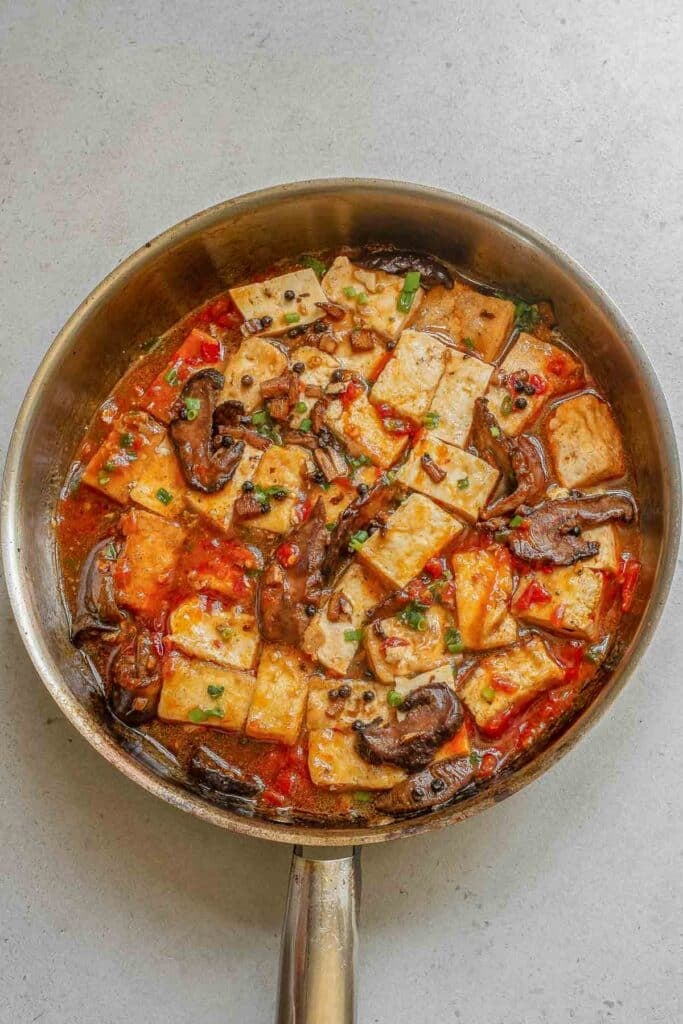 A stainless steel pan filled with tofu, mushrooms in a red sauce, garnished with chopped green onion.