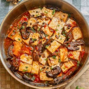 A pan filled with tofu cubes, sliced mushrooms, chopped green onions, and red sauce, garnished with herbs, sits on a woven placemat with ginger in the background.