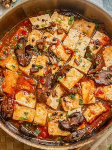 A pan filled with tofu cubes, sliced mushrooms, chopped green onions, and red sauce, garnished with herbs, sits on a woven placemat with ginger in the background.