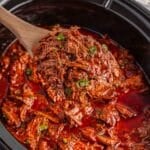 A wooden spoon lifts shredded birria in a thick, red sauce from a slow cooker, garnished with chopped herbs.