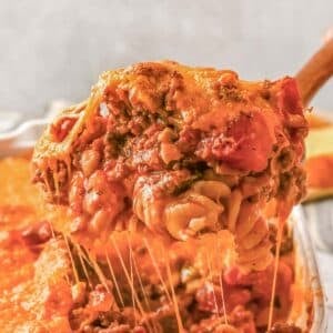 A close-up of a serving of cheesy baked Johnny Marzetti recipe with ground meat and tomato sauce being lifted from a casserole dish.