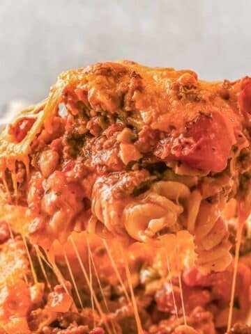 A close-up of a serving of cheesy baked Johnny Marzetti recipe with ground meat and tomato sauce being lifted from a casserole dish.