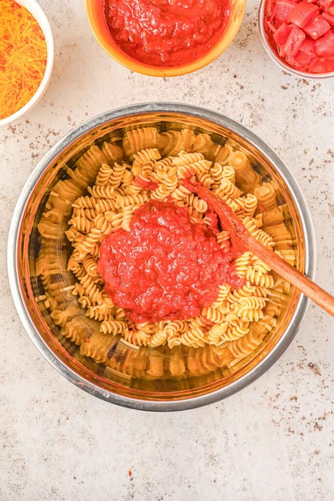 A metal bowl of cooked rotini pasta with tomato sauce on top, next to bowls of shredded cheese, diced tomatoes, and extra sauce.