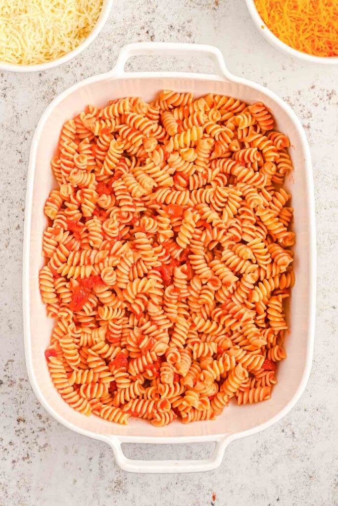 Rectangular white baking dish filled with rotini pasta in red tomato sauce, with bowls of shredded cheese visible in the background.