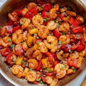 A skillet filled with cooked thai shrimp, red bell peppers, pineapple chunks, and chopped green onions in a glossy sauce.