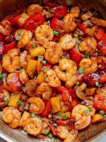 A skillet filled with cooked thai shrimp, red bell peppers, pineapple chunks, and chopped green onions in a glossy sauce.