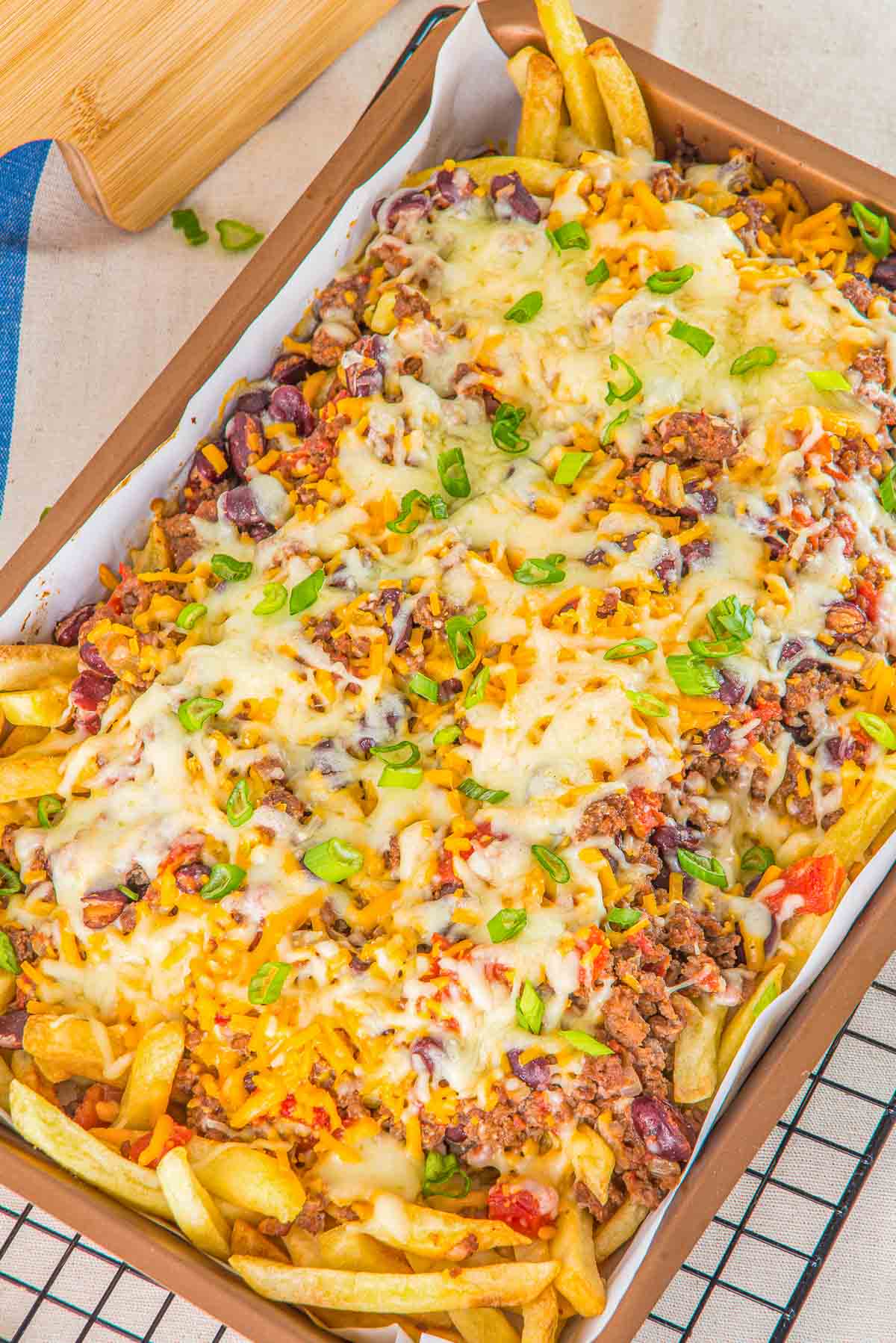 A tray of chili cheese fries topped with melted cheese, ground meat, kidney beans, corn, chopped tomatoes, and sliced green onions on a metal wire rack.