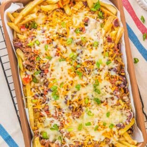A tray of chili cheese fries topped with melted cheese, ground meat, kidney beans, corn, chopped tomatoes, and sliced green onions on a metal wire rack.