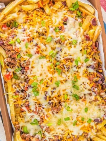 A tray of chili cheese fries topped with melted cheese, ground meat, kidney beans, corn, chopped tomatoes, and sliced green onions on a metal wire rack.