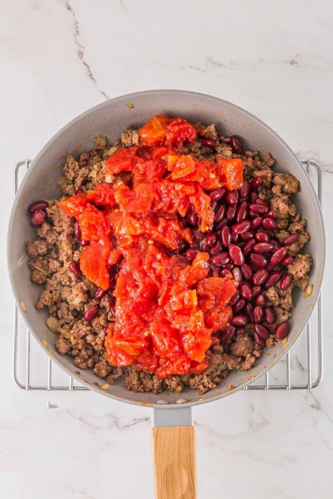 A frying pan containing cooked ground meat, kidney beans, and diced tomatoes, placed on a cooling rack over a marble surface.