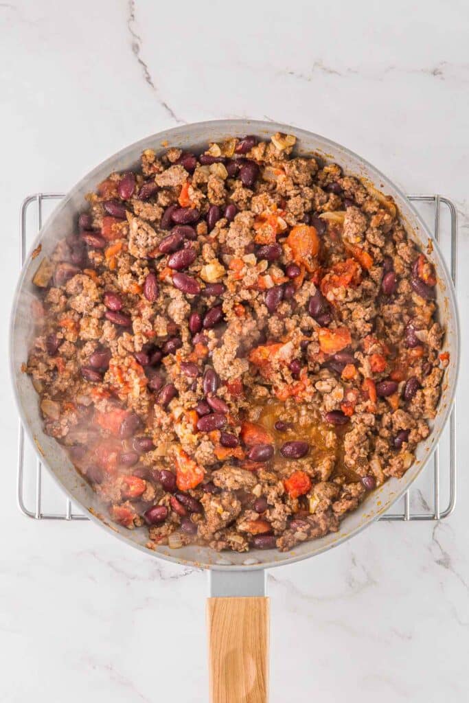 A skillet filled with cooked ground beef, kidney beans, and diced tomatoes with spices set on a wire rack.
