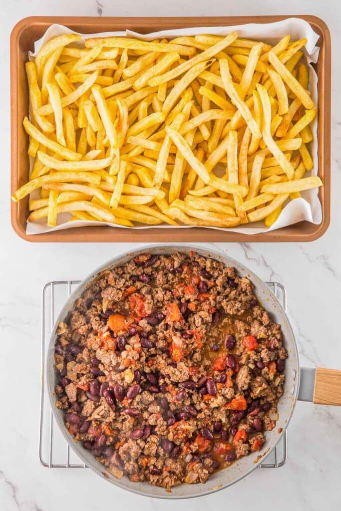A baking tray of uncooked French fries with a skillet of chili off to the side.