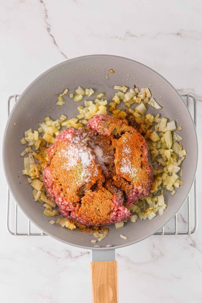 Ground beef with seasonings and chopped onions cooking in a skillet.