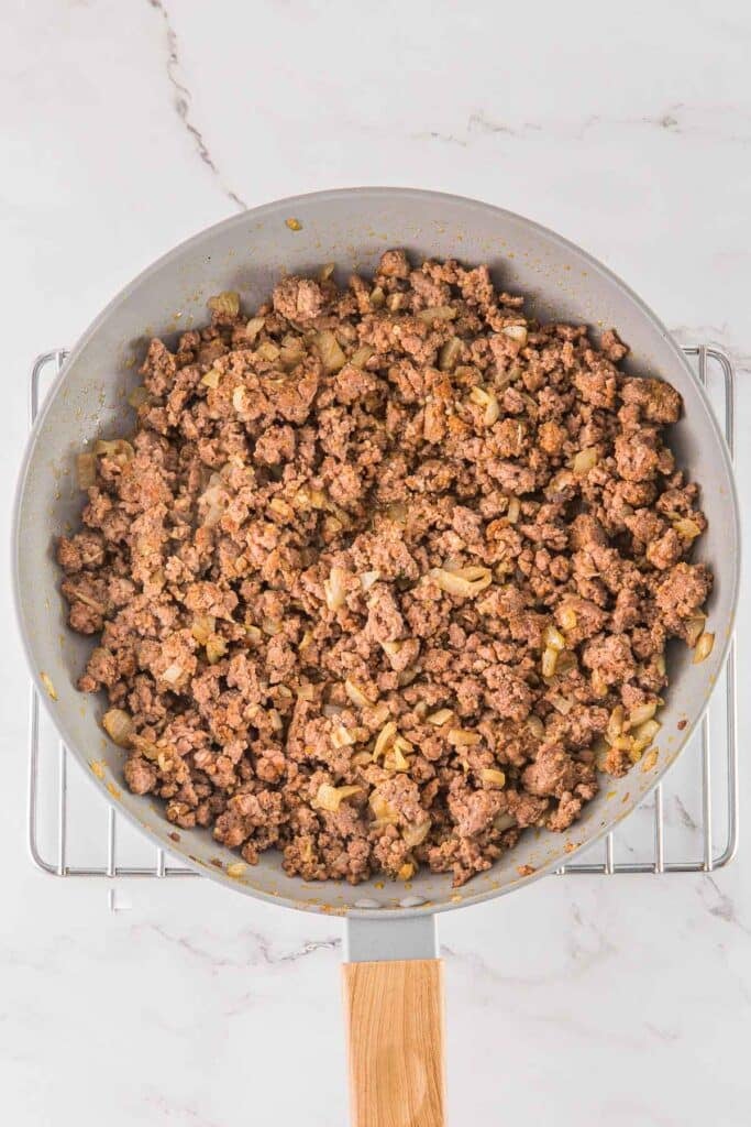 Cooked ground beef with chopped onions in a frying pan, placed on a metal rack over a marble countertop.