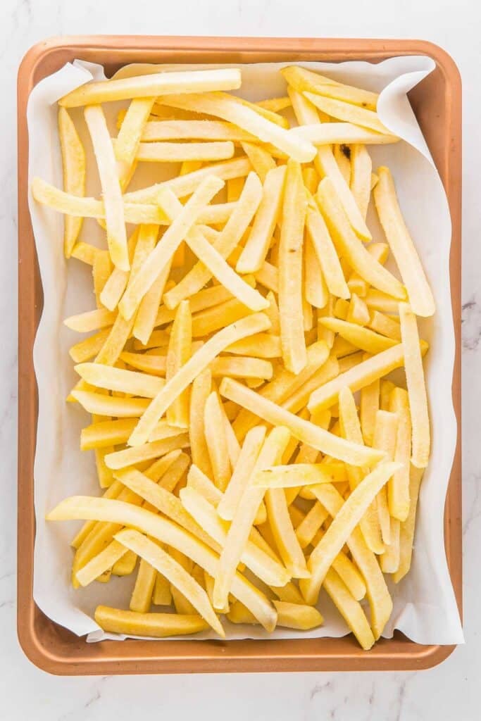 A baking sheet lined with parchment paper is filled with uncooked frozen French fries.