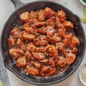 A cast iron skillet filled with empress chicken garnished with chopped green onions and sesame seeds.