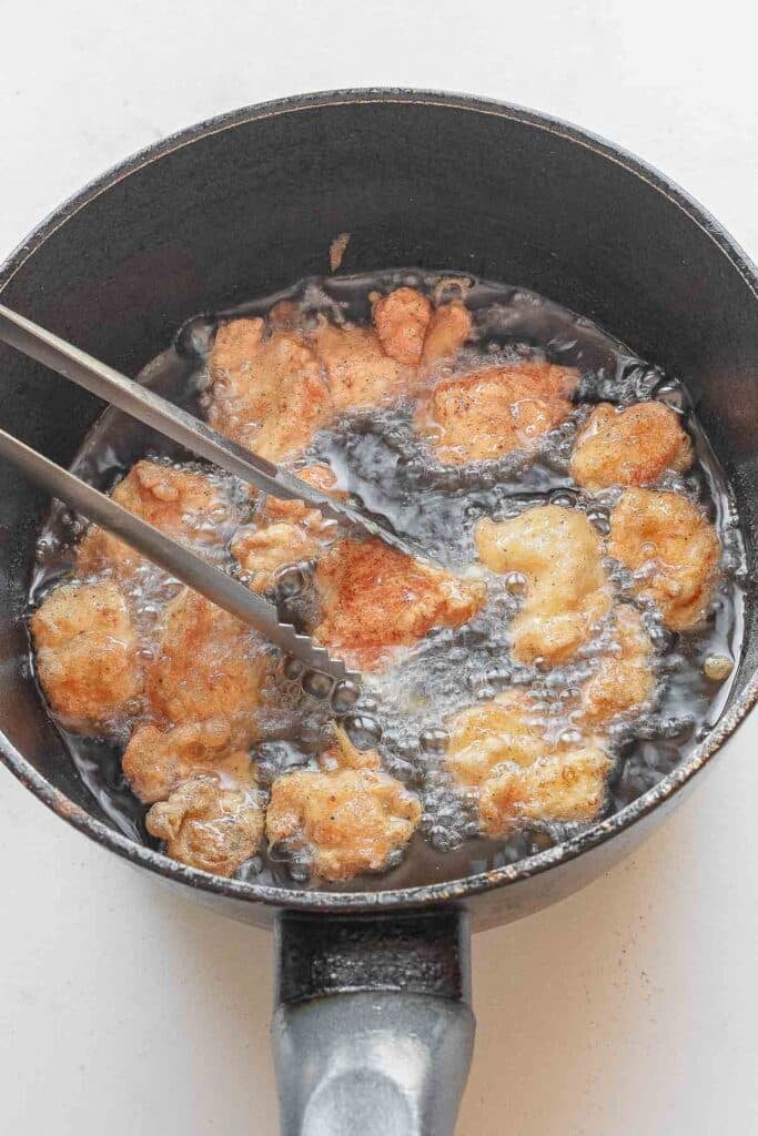 Chicken pieces being fried in hot oil in a black pan, with metal tongs holding one piece.