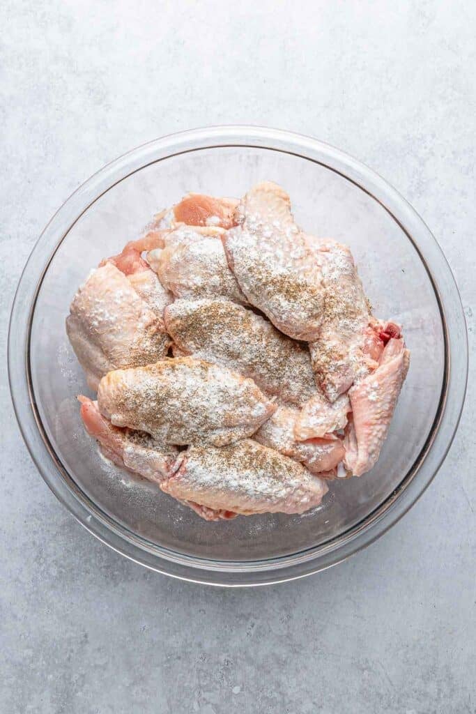 Raw chicken wings in a clear glass bowl, coated with baking powder, salt, and pepper.
