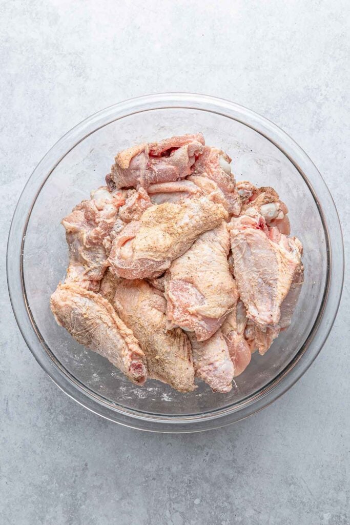 Raw chicken wings in a clear glass bowl, tossed with baking powder, salt, and pepper.