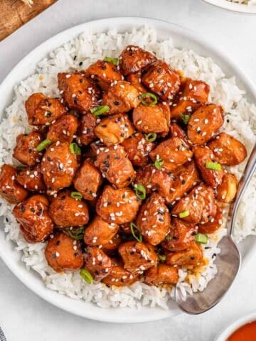 Bowl of white rice topped with honey garlic chicken pieces, garnished with sesame seeds and green onions.