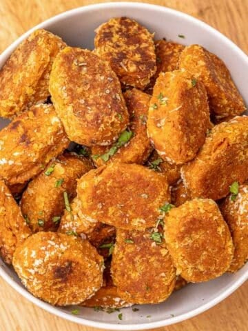 A white bowl filled with sweet potato tater tots garnished with chopped herbs on a wooden cutting board.