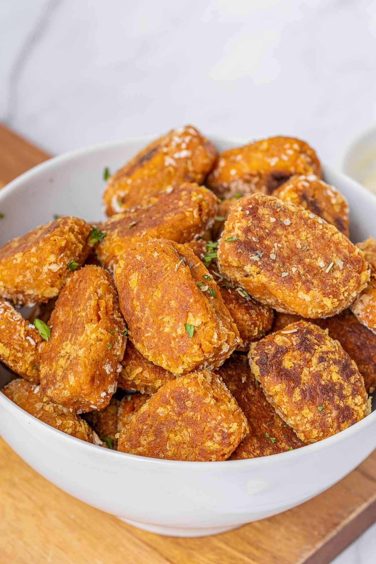 A white bowl filled with sweet potato tater tots garnished with chopped herbs, placed on a wooden surface.