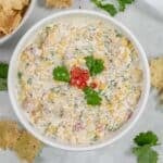 A white bowl filled with an easy cowboy corn dip recipe, garnished with cilantro and diced tomatoes, surrounded by tortilla chips.