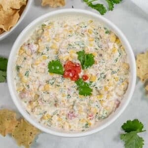 A white bowl filled with an easy cowboy corn dip recipe, garnished with cilantro and diced tomatoes, surrounded by tortilla chips.