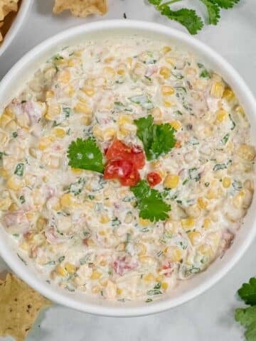 A white bowl filled with an easy cowboy corn dip recipe, garnished with cilantro and diced tomatoes, surrounded by tortilla chips.
