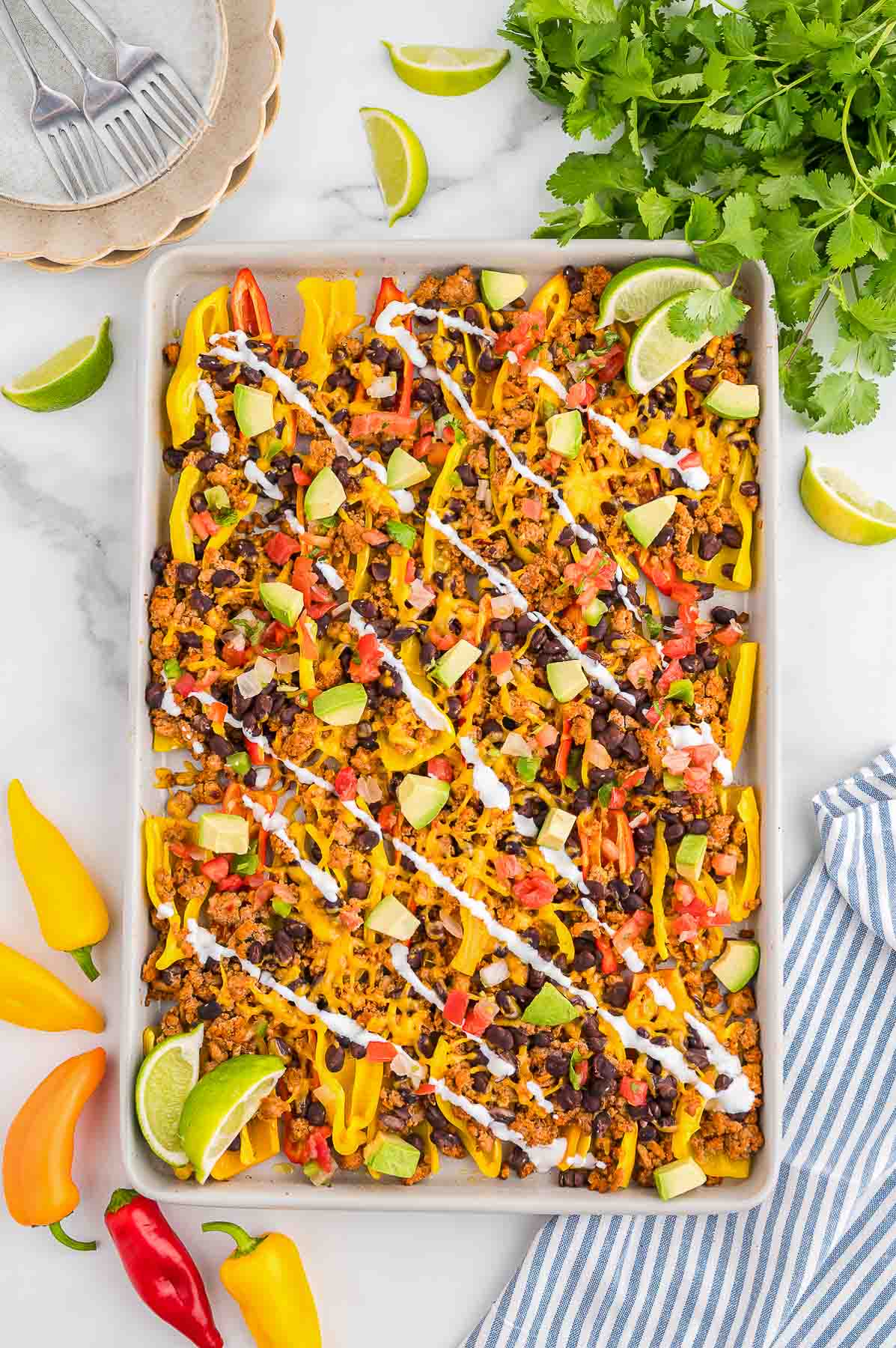A sheet pan filled with bell pepper nachos topped with ground meat, black beans, diced avocado, tomatoes, cheese, and drizzled with white sauce, garnished with lime wedges.