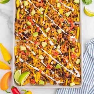 A sheet pan filled with bell pepper nachos topped with ground meat, black beans, diced avocado, tomatoes, cheese, and drizzled with white sauce, garnished with lime wedges.