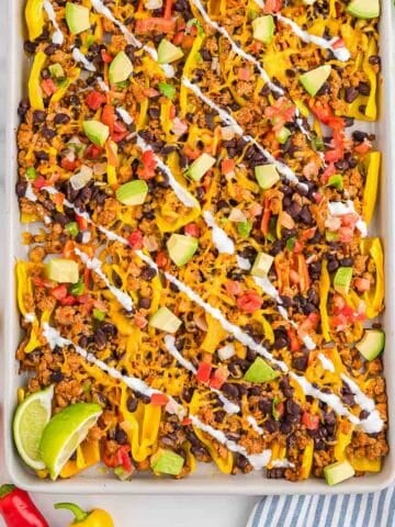 A sheet pan filled with bell pepper nachos topped with ground meat, black beans, diced avocado, tomatoes, cheese, and drizzled with white sauce, garnished with lime wedges.