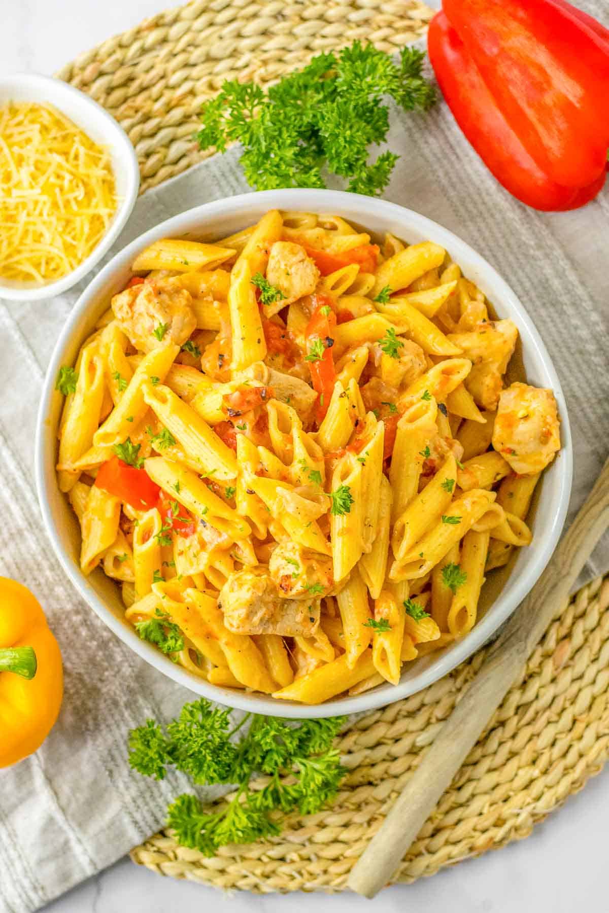 A bowl of cajun chicken pasta with chicken, bell peppers, and herbs is placed on a woven mat, next to a small bowl of shredded cheese, parsley, and a sliced red bell pepper.