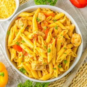 A bowl of cajun chicken pasta with chicken, bell peppers, and herbs is placed on a woven mat, next to a small bowl of shredded cheese, parsley, and a sliced red bell pepper.
