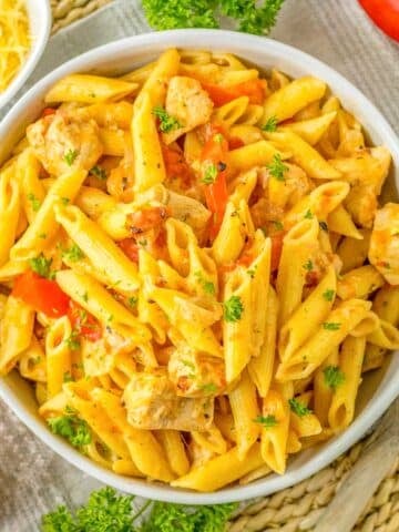 A bowl of cajun chicken pasta with chicken, bell peppers, and herbs is placed on a woven mat, next to a small bowl of shredded cheese, parsley, and a sliced red bell pepper.