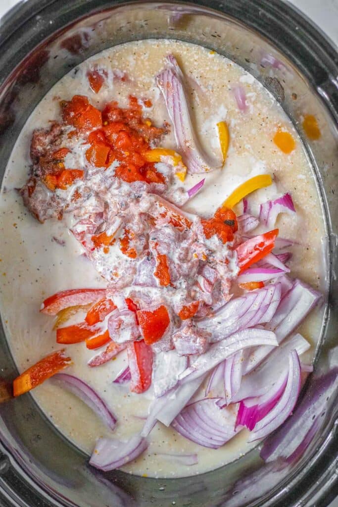 Sliced red onions, bell peppers, diced tomatoes, chicken broth and heavy cream in a slow cooker, ready to be cooked.