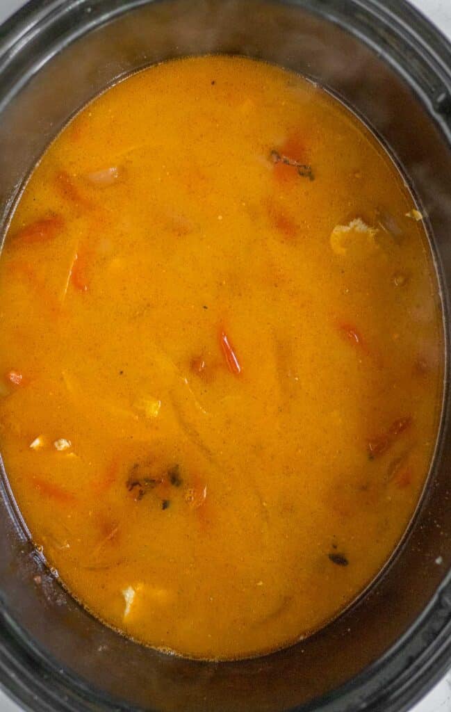 A close-up of a pot filled with orange-colored soup or stew, containing visible pieces of vegetables and herbs.