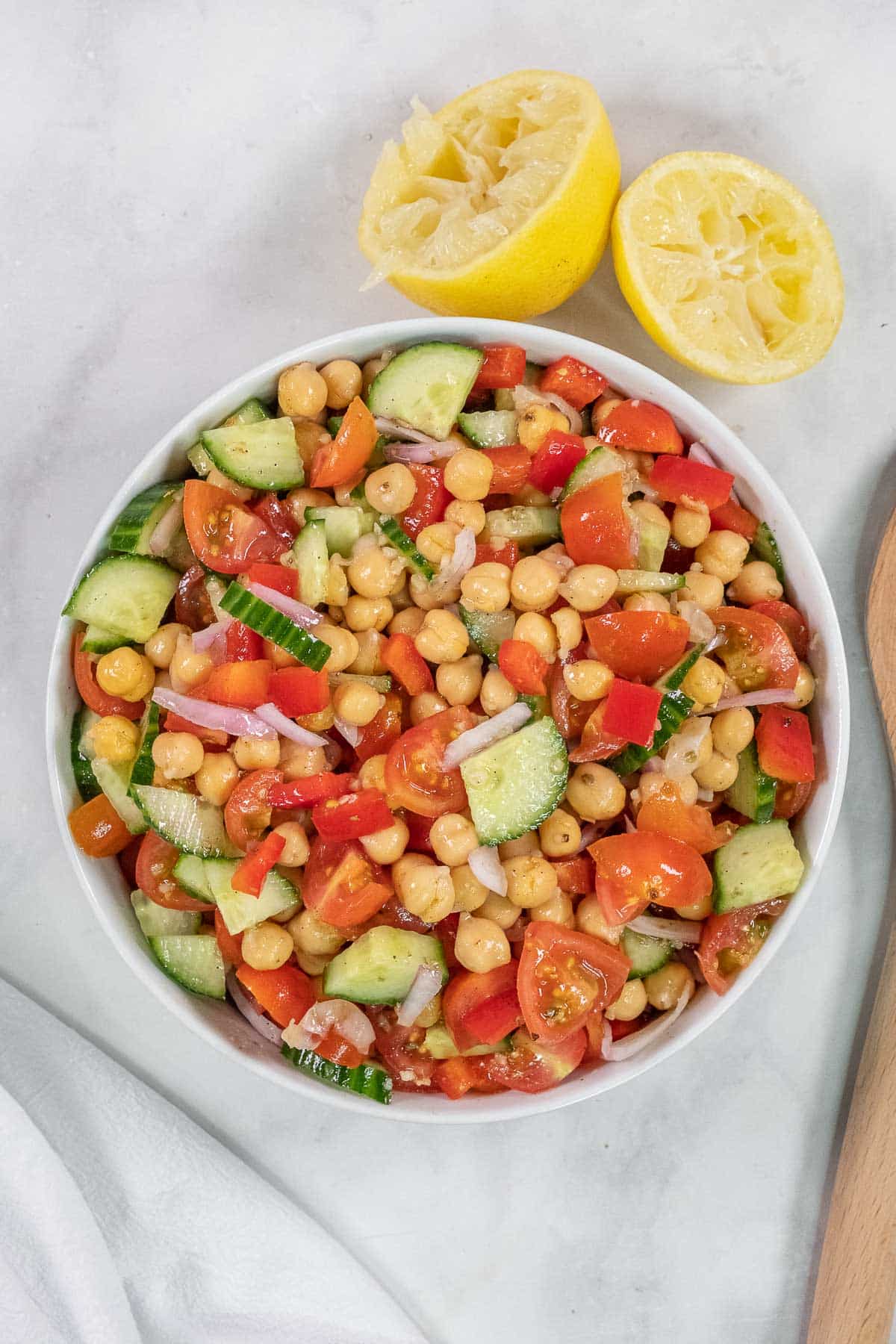 A bowl of chickpea salad with cucumbers, cherry tomatoes, red onions, and red bell peppers, next to a halved, squeezed lemon on a white surface.