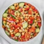 A bowl of chickpea salad with chopped cucumbers, tomatoes, red bell peppers, and red onions on a white surface.