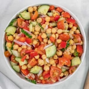 A bowl of chickpea salad with chopped cucumbers, tomatoes, red bell peppers, and red onions on a white surface.