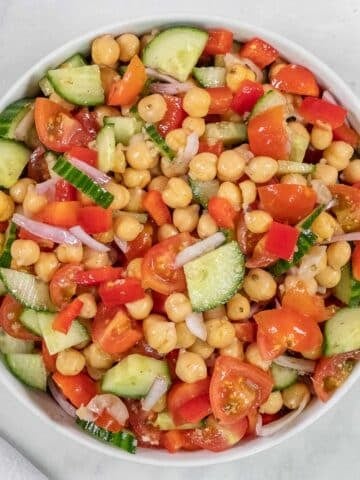 A bowl of chickpea salad with chopped cucumbers, tomatoes, red bell peppers, and red onions on a white surface.