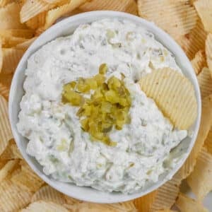 A bowl of creamy dill pickle dip topped with chopped pickles with one chip dipped in the dip, surrounded by potato chips on a white plate.