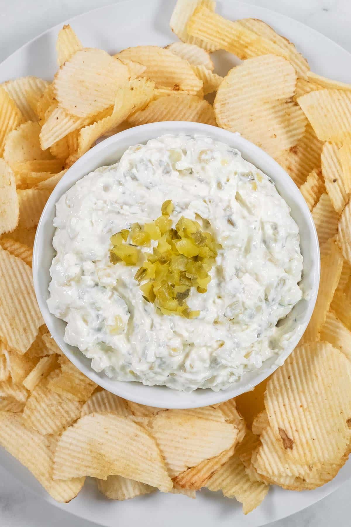 A bowl of creamy dill pickle dip topped with chopped pickles, surrounded by potato chips on a white plate.