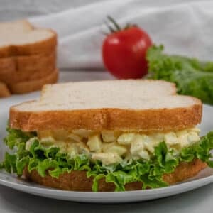 Egg salad sandwich with leaf lettuce on white bread, served on a white plate. A tomato, lettuce, and sliced bread are in the background.