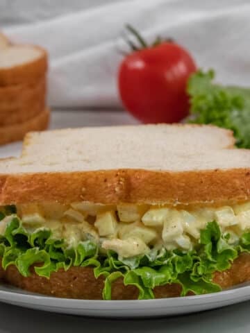 Egg salad sandwich with leaf lettuce on white bread, served on a white plate. A tomato, lettuce, and sliced bread are in the background.