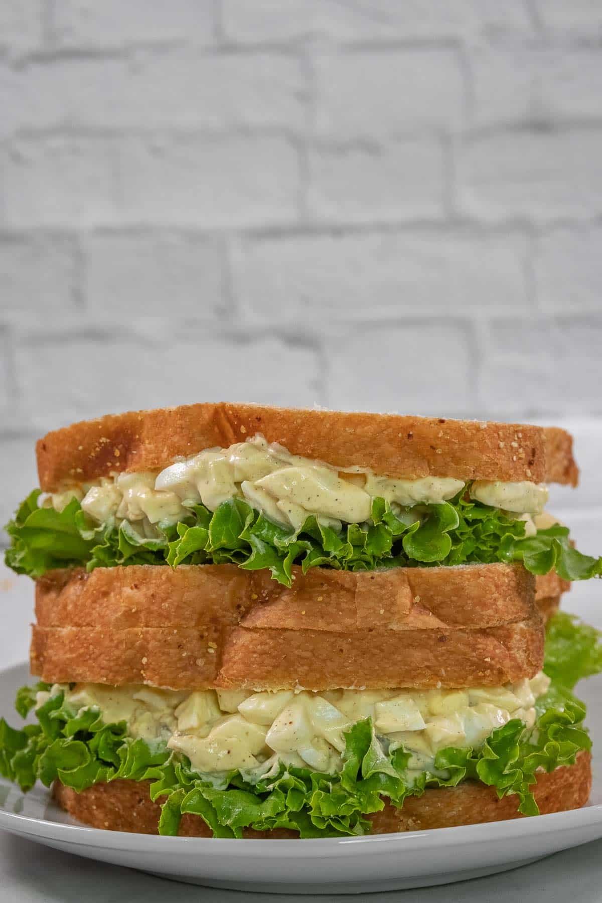 Two stacked egg salad sandwiches with lettuce on white bread, served on a white plate against a light brick background.