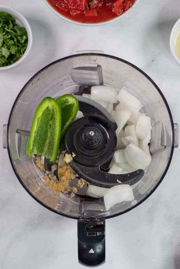 A food processor filled with chopped white onion, halved jalapeno pepper, and minced garlic, viewed from above.