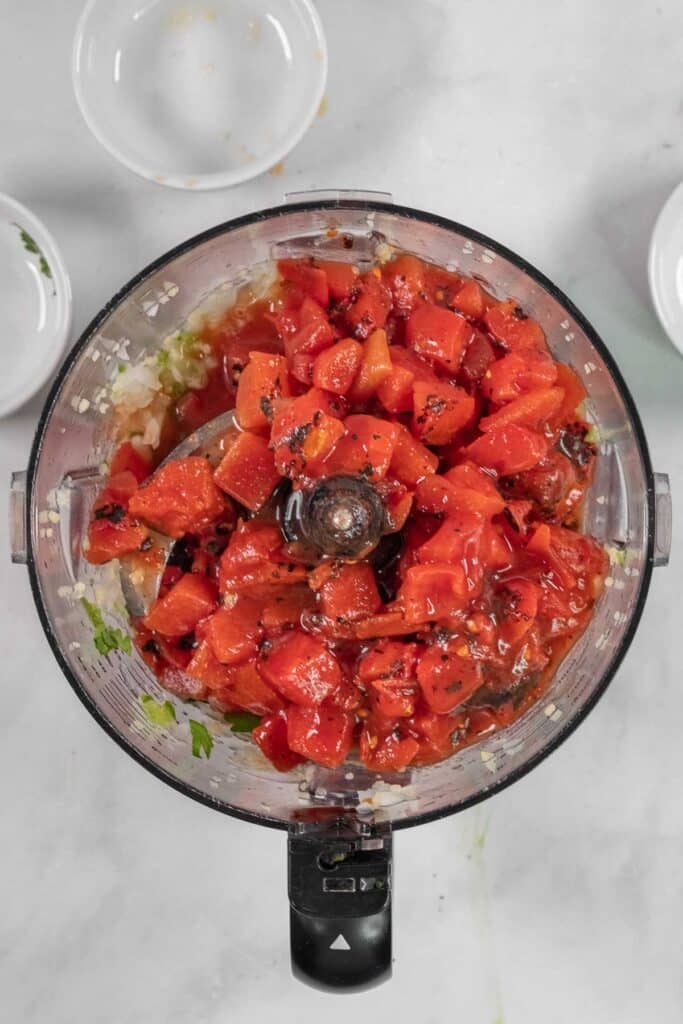 A food processor with chopped tomatoes added in with the rest of the mixture onion and pepper mixture.