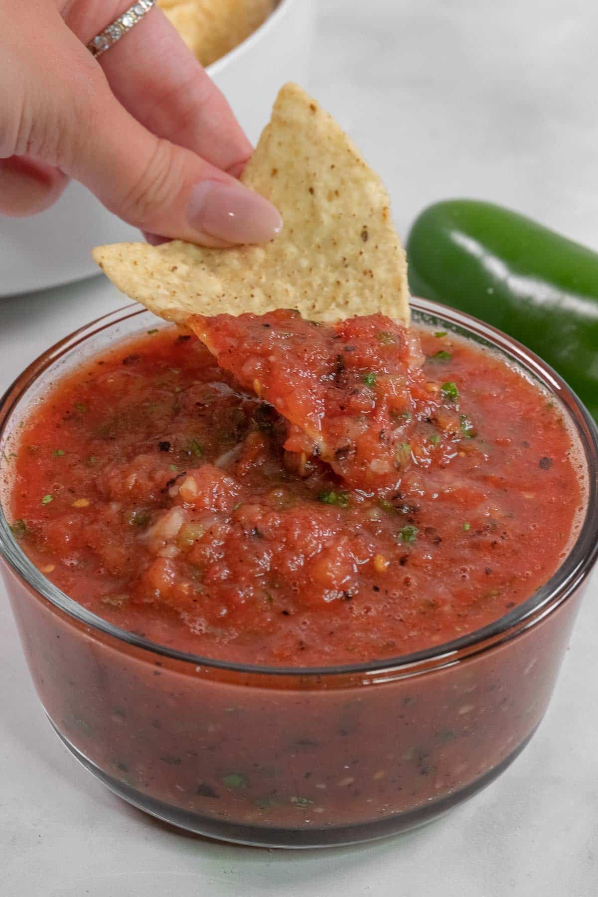 A hand dips a tortilla chip into a glass bowl of red salsa, with a green jalapeño pepper.